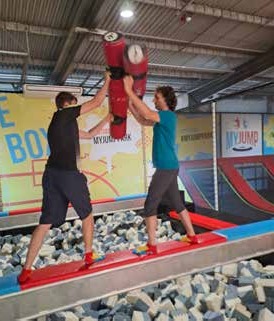 In einer Trampolinhalle hebt ein junger Mann einen anderen hoch, der Boden ist mit grauen Schaumstoffwürfeln gefüllt.