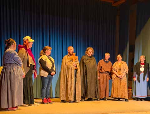 Die Theatergruppe steht in mittelalterlichen Kostümen – Umhänge, Kopftücher und Kutten – nebeneinander auf der Bühne vor einem blauen Vorhang.