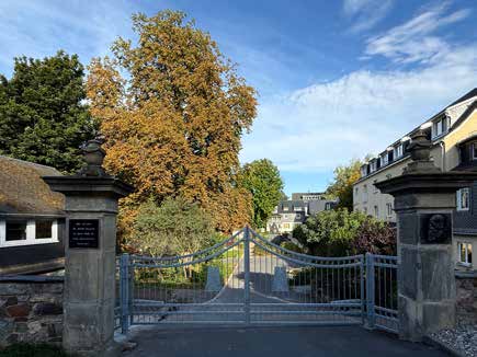 Das Eingangstor der Lebensgemeinschaft zwischen zwei steinernen Pfeilern, dahinter ein Baum in Herbstfarben und die Häuser im Sonnenlicht.