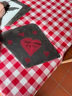 Auf einer Schiefertafel ist ein rotes Herz mit Schrift gemalt, daneben kleine Herzen. Die Tafel liegt auf einer rot-weiß karierten Tischdecke.