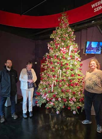 Drei Ausflügler stehen neben einem großen, reich geschmückten Weihnachtsbaum mit roten und weißen Kugeln.
