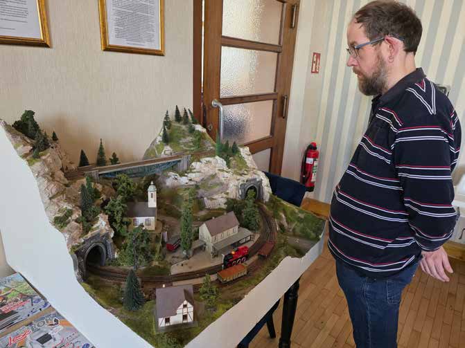 Ein Bewohner mit Brille betrachtet eine große Modelleisenbahn-Landschaft mit Bergen, Tunneln und einer kleinen Kirche.