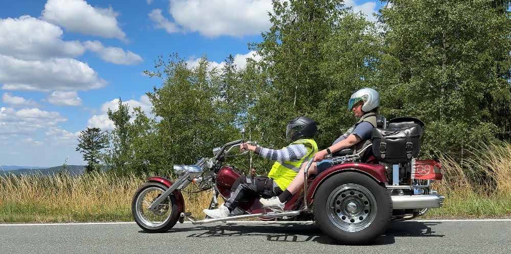 Ein dunkelrotes Trike mit zwei Personen fährt auf einer Landstraße, im Hintergrund Wiesen, Bäume und Wolken.
