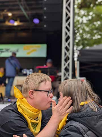 Ein junger Mann mit gelbem Halstuch und Brille berührt zärtlich das Gesicht einer Frau, im Hintergrund die Bühne.