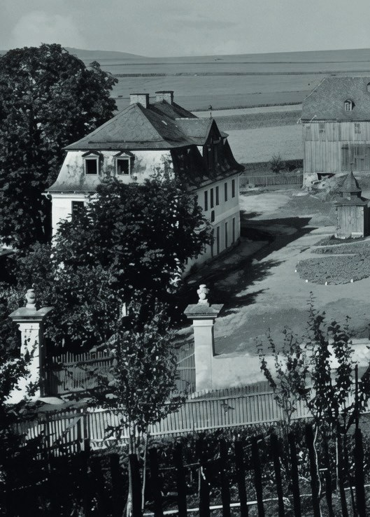Historisches Schwarz-Weiß-Foto: Ein stattliches Herrenhaus mit Mansarddach hinter einem Tor mit steinernen Pfeilern, dahinter Felder.