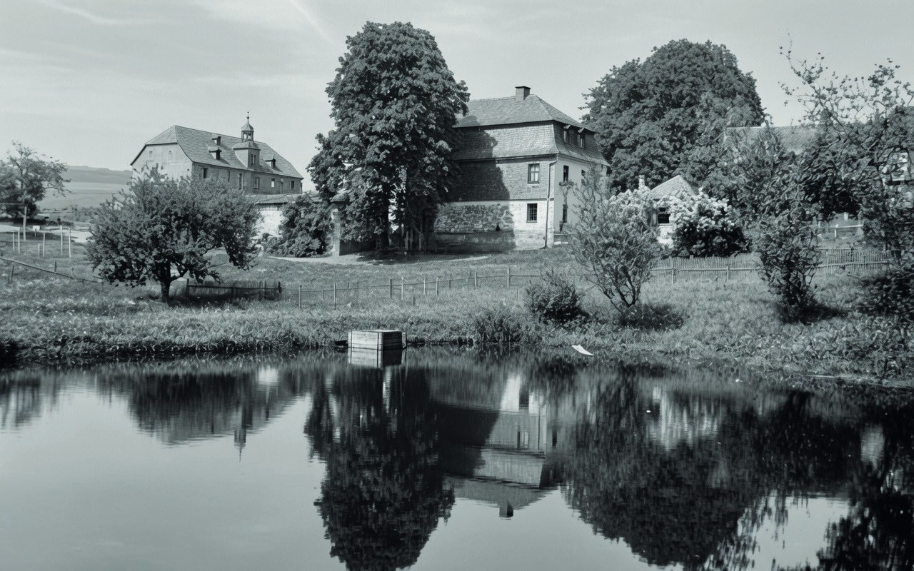 Historisches Schwarz-Weiß-Foto: Ein Teich spiegelt Bäume und Gebäude des Guts, darunter ein Haus mit Türmchen.