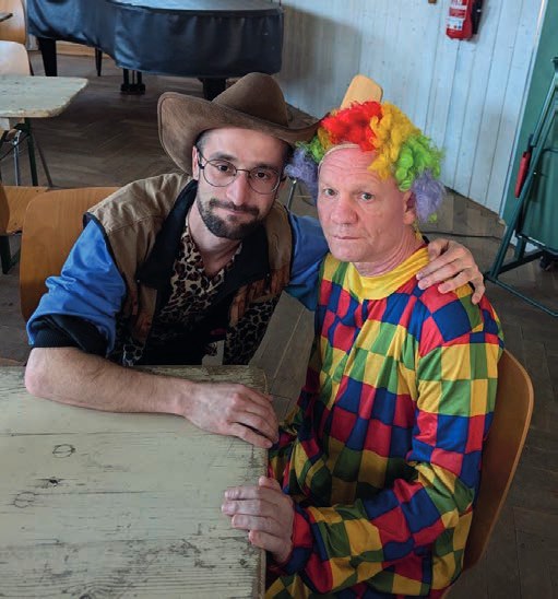 Ruslan Solntsev mit Cowboyhut und ein Bewohner in buntem Clownskostüm mit Regenbogenperücke sitzen nebeneinander an einem Tisch.